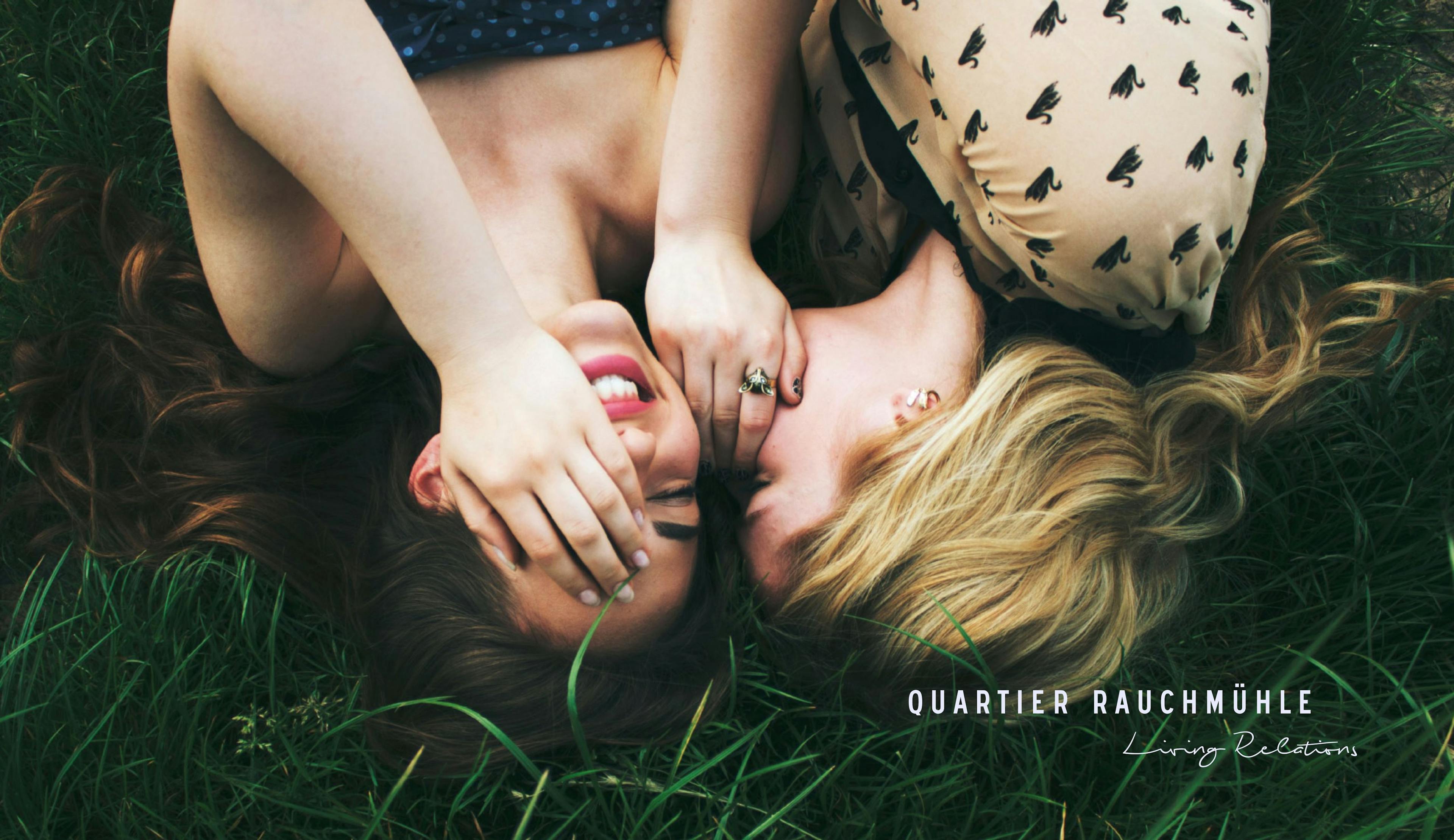 Zwei jungen Frauen, die in der Wiese liegen und sich etwas ins Ohr flüstern. © gobiq